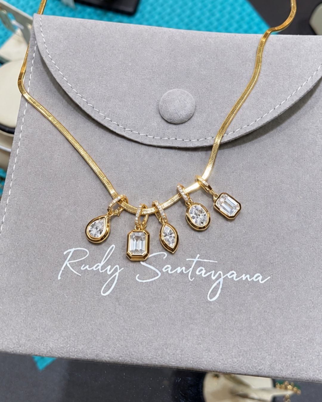 A gold necklace with five dangling diamond charm pendants, displayed on a gray velvet jewelry pouch with the brand name Rudy Santayana printed on it.